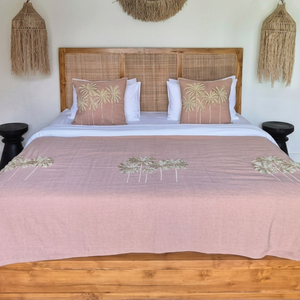 A styled bedroom scene featuring a wooden bed frame with a woven headboard, dressed in a dusty pink tropical-print blanket and matching pillows, set against a light cream wall with bohemian wall decor.