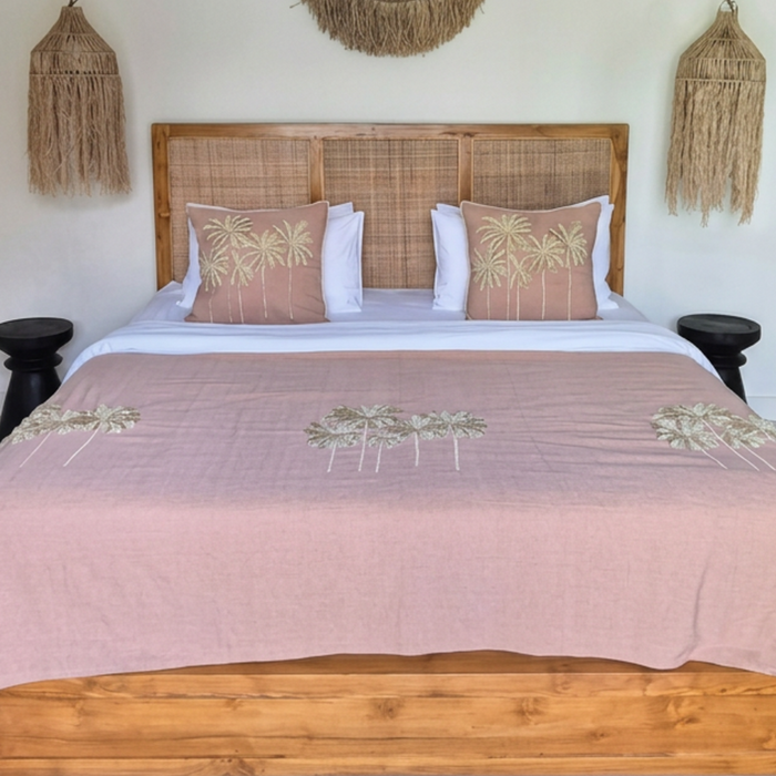 A styled bedroom scene featuring a wooden bed frame with a woven headboard, dressed in a dusty pink tropical-print blanket and matching pillows, set against a light cream wall with bohemian wall decor.