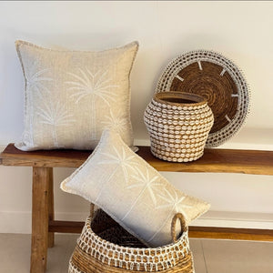 Two beige linen throw pillows with white palm tree embroidery are styled on and around a rustic wooden bench with woven baskets against a white wall.