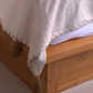 A close-up of a light beige linen throw blanket with decorative tassels and a stitched border, draped over a wooden bed frame.