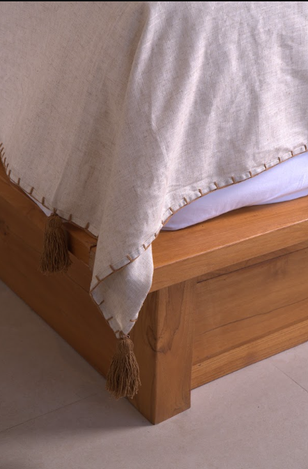 A close-up of a light beige linen throw blanket with decorative tassels and a stitched border, draped over a wooden bed frame.