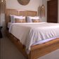 A wooden platform bed with a woven cane headboard, dressed in white linens and two beige accent pillows with a palm tree motif, set in a minimalist bedroom with boho-style woven pendant lights.