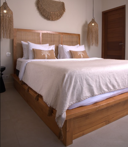 A wooden platform bed with a woven cane headboard, dressed in white linens and two beige accent pillows with a palm tree motif, set in a minimalist bedroom with boho-style woven pendant lights.