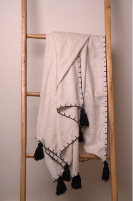 A linen cotton blend natural throw blanket with black tassels and a black geometric border pattern, draped over a wooden ladder