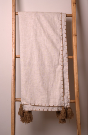 A light beige linen and cotton blend  throw blanket with brown tassels and a decorative stitched border, draped over a wooden ladder.