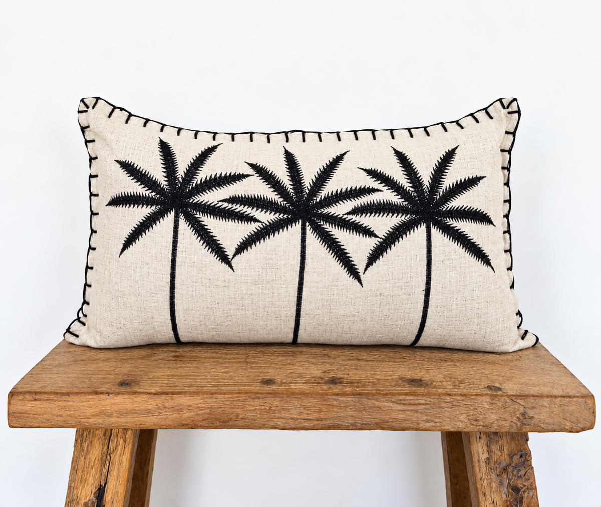 A rectangular beige linen throw pillow with black embroidered palm tree silhouettes and black whipstitching along the edges, displayed on a rustic wooden bench against a white background.