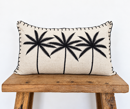 A rectangular beige linen throw pillow with black embroidered palm tree silhouettes and black whipstitching along the edges, displayed on a rustic wooden bench against a white background.