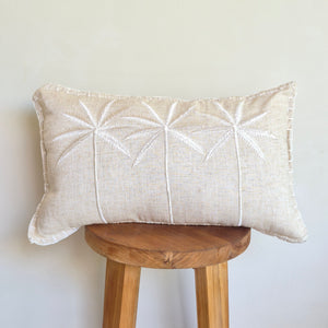 A rectangular cream linen accent pillow with white embroidered palm trees patterns, resting on a wooden stool.
