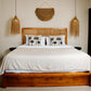A rustic wooden platform bed with white bedding and two black palm tree patterned pillows, styled in a minimalist bedroom with a woven wall hanging and two hanging woven lamps.
