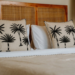 A close-up of a bed with beige linens, white pillows, and two decorative throw pillows featuring black palm tree prints, set against a wooden headboard with woven cane panels.