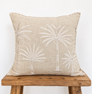 A square beige linen throw pillow with white embroidered palm tree motifs.