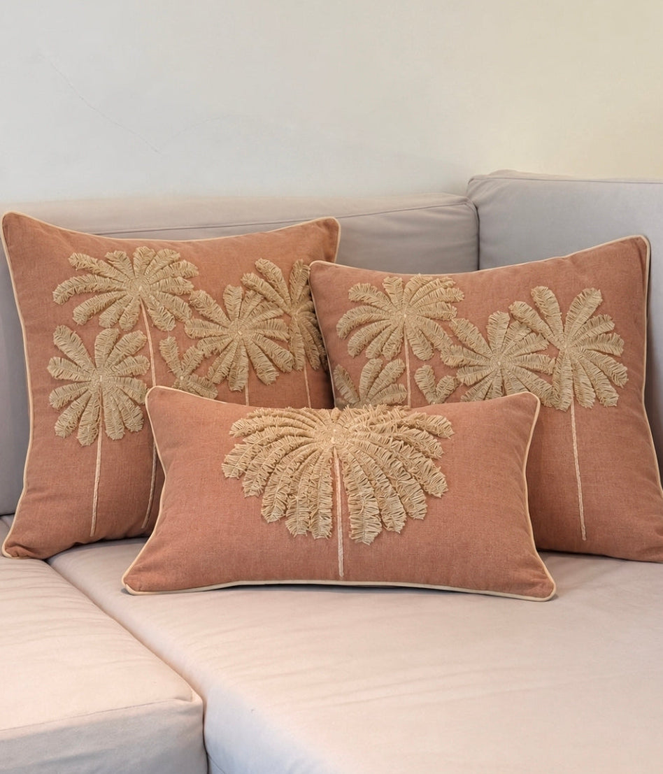 Three rectangular throw pillows in a dusty terracotta pink color with beige palm tree embroidery, arranged on a light gray sofa.