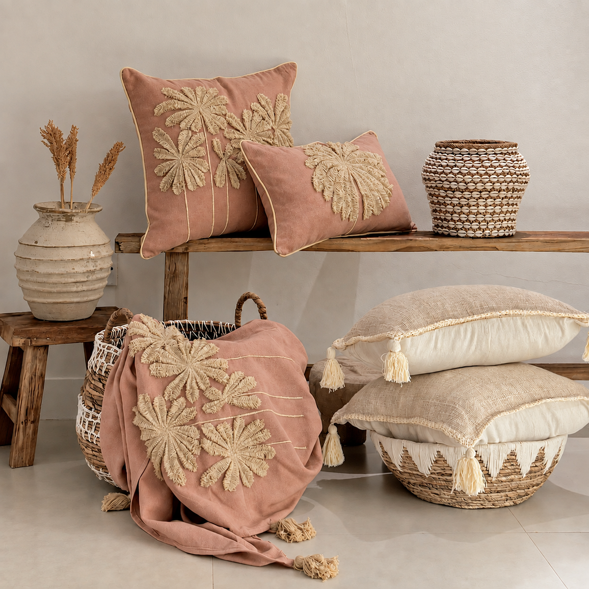 A styled home decor arrangement featuring dusty pink throw pillows with large beige floral embroidery, a matching pink throw blanket draped over a woven basket, and additional neutral-toned cushions and baskets on a wooden bench and floor against a light beige wall.