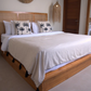 A wooden platform bed with a woven headboard, dressed in white bedding and two black palm tree patterned accent pillows, set in a minimalist bedroom with a black side table and a mirror.