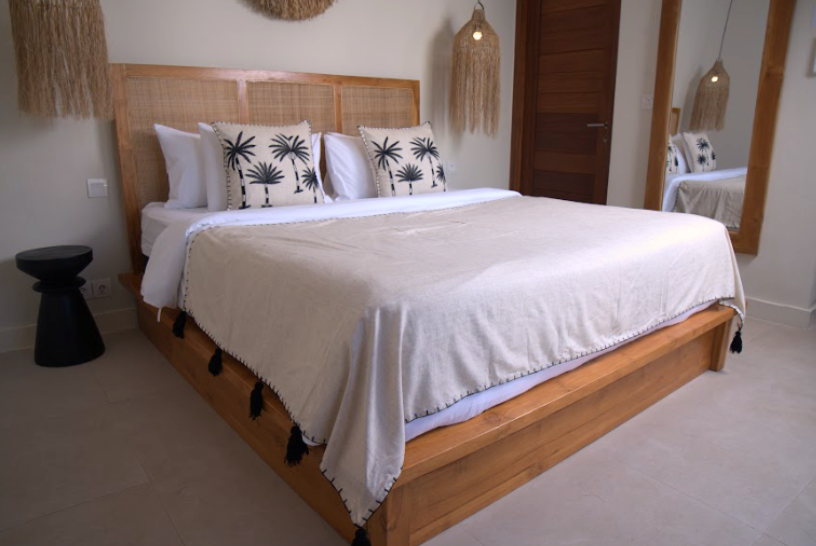 A wooden platform bed with a woven headboard, dressed in white bedding and two black palm tree patterned accent pillows, set in a minimalist bedroom with a black side table and a mirror.