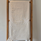 A light beige linen and cotton blen throwr with white embroidered palm tree details and white fringe tassels, displayed on a rustic wooden rack.