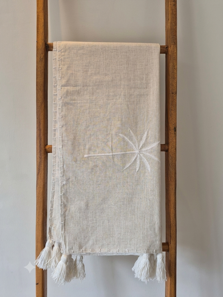 A light beige linen and cotton blen throwr with white embroidered palm tree details and white fringe tassels, displayed on a rustic wooden rack.